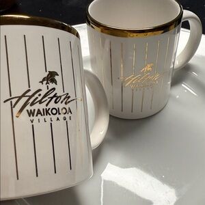 ceramic mugs feature gold-toned vertical stripes 
Hilton Waikoloa Village logo